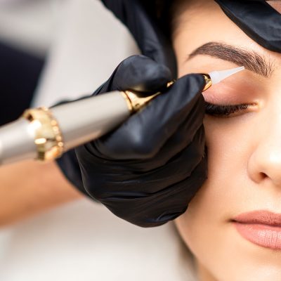 Beautician applying permanent makeup on eyebrows of young woman by special tattoo machine tool